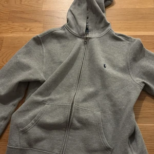 Grå hoodie från Ralph Lauren - Säljer en klassisk grå hoodie från Ralph Lauren med dragkedja och fickor framtill. Perfekt för en avslappnad stil. Den har en liten logga broderad på bröstet och en bekväm huva.