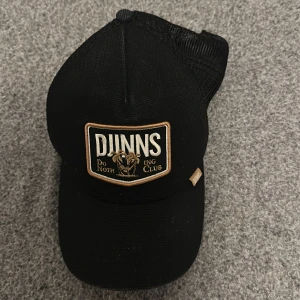 Svart keps från Djinns - Snygg svart keps från Djinns med ett broderat märke framtill som säger 'Do Nothing Club'. Kepsen har ett justerbart spänne bak och meshmaterial för extra ventilation. Perfekt för en avslappnad stil.