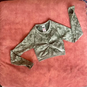 Grön camo-mönstrad croppad tröja - Snygg grön camo-mönstrad croppad tröja från Gymshark. Perfekt för träning med långärmad design och en stilren öppning i ryggen. Materialet är stretchigt och bekvämt, vilket gör den idealisk för aktiva dagar.