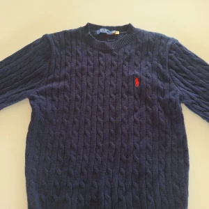 Mörkblå stickad tröja från Polo Ralph Lauren - Säljer en stilren mörkblå stickad tröja från Polo Ralph Lauren med kabelstickat mönster och röd logga på bröstet. Tröjan har långa ärmar och rund halsringning. Perfekt för kyliga dagar!