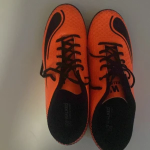 Orange fotbollsskor med svarta detaljer - Snygga orange fotbollsskor med svarta detaljer och snörning. Perfekta för spel på gräs med sin greppvänliga sula. Skorna har en modern design med en tydlig logotyp på sidan.