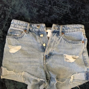 Blå jeansshorts från H&M - Säljer ett par blå jeansshorts från H&M i storlek 36. De har en högmidjad passform med slitna detaljer och uppvikta kanter. Perfekta för en avslappnad stil.