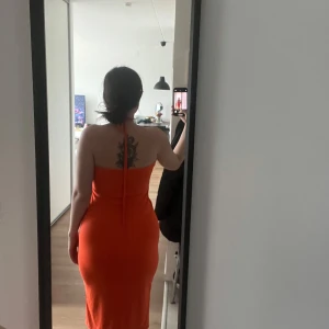 Orange klänning med halterneck - Snygg orange klänning med halterneck-design och en unik detalj framtill. Klänningen har en tight passform och är perfekt för sommarens fester. Den är ärmlös och har en smickrande längd.