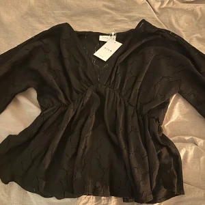 Svart blus från Vila - Säljer en elegant svart blus från Vila med v-ringning och långa ärmar. Blusen har ett subtilt mönster och en lätt volang vid nederkanten. Perfekt för en stilren look. Nypris 529 kr