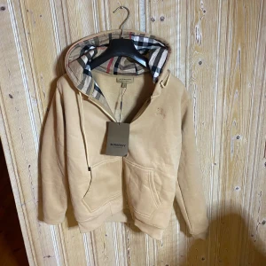 Burberry kofta - Säljer en snygg beige kofta från Burberry med klassiskt rutigt mönster i luvan. Tröjan har en dragkedja framtill och en praktisk ficka. Perfekt för en stilren och avslappnad look.
