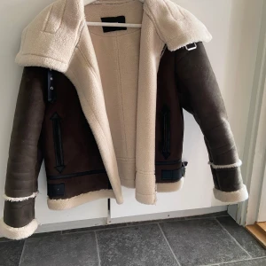 Brun och beige jacka från Weekday - Säljer en snygg brun och beige jacka från Weekday. Jackan har en mysig pälsfoder och dragkedja framtill. Den har långa ärmar och detaljer som bälte och spännen vid midjan. Perfekt för kyliga dagar!