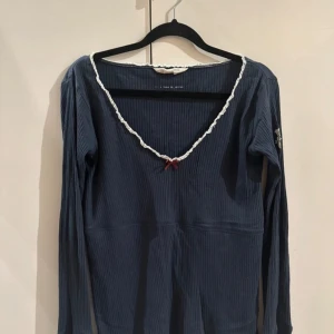 Blå ribbad tröja med rosett - Säljer en blå ribbad tröja från Odd Molly. Tröjan har en v-ringad hals med vit spetskant och en liten röd rosett framtill. Den har långa ärmar och ett broderat emblem på ena ärmen.