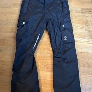 Svarta täckbyxor från Dope - Snygga svarta täckbyxor från Dope med praktiska fickor och dragkedjor. Perfekta för vinterns äventyr med en bekväm passform och slitstarkt material. Byxorna har en cool design med orange foder som ger en extra touch.