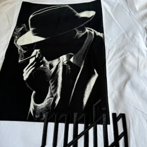 Vit t-shirt med tryck från Ganos - Snygg oversized vit t-shirt från GANGS med ett coolt svartvitt tryck av en person i hatt och texten 'mafia' broderad på baksidan. Perfekt för en stilren och edgy look. Perfekt till gymmet, för någon som gillar att träna i oversized t-shirt. 