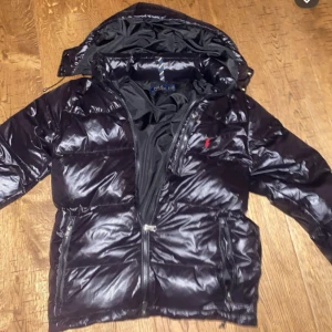 Svart pufferjacka från Polo Ralph Lauren - Säljer en snygg svart pufferjacka från Polo Ralph Lauren. Jackan har en glansig finish med dragkedja och huva. Perfekt för kyliga dagar. Den har långa ärmar och två praktiska fickor med dragkedja framtill.
