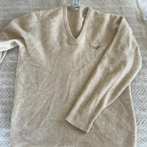 Beige v-ringad tröja  - Säljer en stilren beige tröja med v-ringning och broderad logga på bröstet. Tröjan har långa ärmar och ribbade muddar vid ärmslut och nederkant. Perfekt för en avslappnad men snygg look.