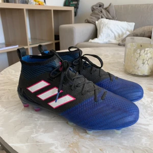 Blå och svarta fotbollsskor från Adidas - Säljer ett par blå och svarta fotbollsskor från Adidas med vita och rosa detaljer. Skorna har snörning och en mönstrad yta för bättre grepp på planen. Perfekta för fotbollsspelare som vill ha stil och funktionalitet. Pris kan diskuteras 