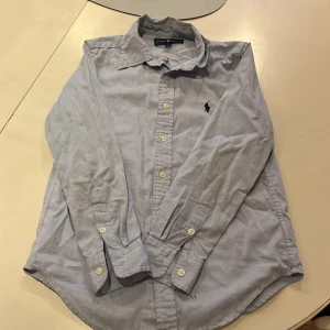 Polo Ralph Lauren skjorta - Säljer en stilren ljusblå skjorta från Ralph Lauren med size 12 år. Skjortan har bra skick det ända är att den är lite skrynklig med det är bara att stryka så blir den bra. Och ett litet mini hål som knappt syns. Ny pris 1000 kr. Skriv vid funderingar! Kan få ner i pris vid snabb affär.
