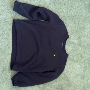 Svart tröja från Lyle & Scott - Säljer en svart tröja från Lyle & Scott med ett gult broderat emblem på bröstet. Tröjan har långa ärmar och en rund halsringning. Perfekt för en stilren och avslappnad look.