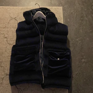 Mörkblå dunväst med dragkedja (limiterad) - Snygg mörkblå dunväst med dragkedja och praktiska fickor. Perfekt för kyligare dagar med sin quiltade design och huva. Västen har en stilren look och passar bra till olika outfits.