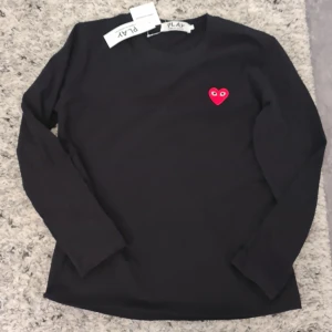 Svart långärmad tröja från Comme des Garçons - Säljer en stilren svart långärmad tröja från Comme des Garçons med det ikoniska röda hjärtat med ögon på bröstet. Perfekt för en minimalistisk look. 💖