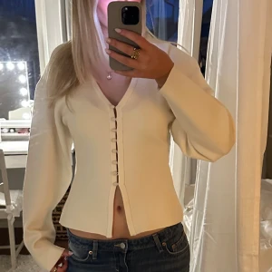 Vit topp med knappar - Säljer en stilren vit kofta med knappar framtill. Koftan har en v-ringad design och långa ärmar, perfekt för en elegant look. Passar bra till både jeans och kjol.