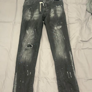 Svarta jeans från Dsquared2 - Snygga svarta jeans från Dsquared2 med coola slitningar och färgstänk i vitt och blått. De har en dragkedja framtill och en tydlig Dsquared2 Ski-logga på baksidan. Perfekta för en trendig look!