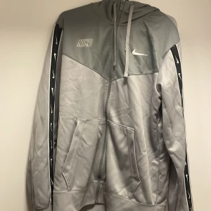 Grå hoodie från Nike - Säljer en grå hoodie från Nike med dragkedja och huva. Jackan har svarta detaljer längs ärmarna med vita Nike-loggor. Perfekt för en sportig look eller avslappnad stil.