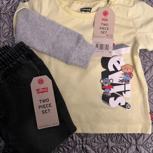 Gul och grå långärmad tröja från Levi's - Säljer en söt gul långärmad tröja från Levi's med grå ärmar och ett tryck av tecknade figurer på framsidan. Perfekt för barn som gillar lekfulla motiv. Tröjan är en del av ett tvådelat set. Jeansbyxor, i mjukt material. Helt nya. 