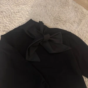 Svart blus med rosett - Elegant svart blus med hög krage och en stor dekorativ rosett vid halsen. Perfekt för en stilren look.