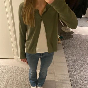Olivgrön kofta med knappar - Säljer en olivgrön kofta med knappar framtill. Den har långa ärmar och en klassisk design som passar perfekt för lager-på-lager-stil. 