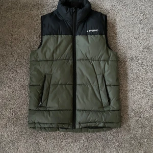 Grön och svart dunväst från Everest - Snygg och praktisk dunväst från Everest i grönt och svart. Västen har dragkedja framtill och två fickor med dragkedjor. Perfekt för lager-på-lager under kyliga dagar och pasar med vilka kläder som helst.