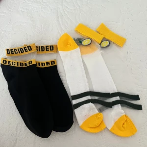 Två par strumpor med text och citronmotiv - Säljer två par strumpor. Ett par är svarta med texten 'DECIDED' i gult på resåren. Det andra paret är vita med gula tår och häl, samt ett citronmotiv vid resåren och svarta ränder runt foten.