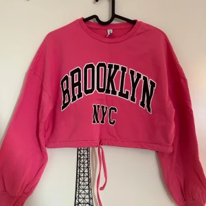 Rosa croppad tröja med Brooklyn-tryck från ONLY  - Rosa croppad tröja med texten 'Brooklyn NYC' i svart och vitt från ONLY i storlek XS. Tröjan har långa ärmar och en justerbar dragsko i nederkanten. Använd några gånger.