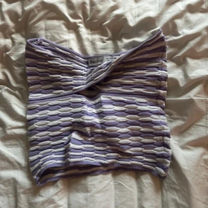 Lila och vit tubtopp från Bershka - Säljer en snygg lila och vit tubtopp från Bershka med ett coolt texturerat mönster. Toppen har en omlott-design framtill som ger en unik look. Perfekt för att styla med dina favoritjeans eller kjol!