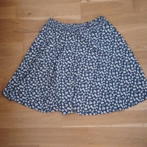 Blommig kjol från Lager 157 - Söt blommig kjol i blått och vitt från lager 157. Kjolen har en elastisk midja för extra komfort och är tillverkad i ett mjukt material med 98% polyester och 2% elastan. Perfekt för en avslappnad stil. Det finns 5 trådar runt om kjolen men 3 trådar har gått av men går fortfarande att använda den och inget av kjolen har gått av pga trådarna. Köpte för 80kr säljer för 35kr.