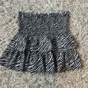 Zebramönstrad kjol från Gina Tricot - Snygg zebramönstrad kjol från Gina Tricot i svart och vitt. Kjolen har en smockad midja och volanger som ger en lekfull look. Perfekt för att sticka ut med stil! Stl 146/152