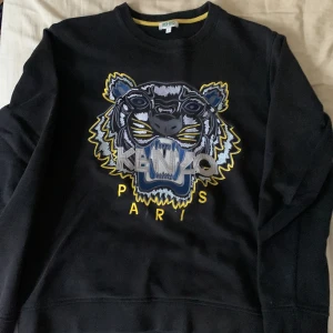 Svart sweatshirt från Kenzo - Säljer en svart sweatshirt från Kenzo med ett stort tigerhuvud broderat på framsidan. Tröjan har långa ärmar och rund halsringning. Perfekt för en stilren och trendig look.