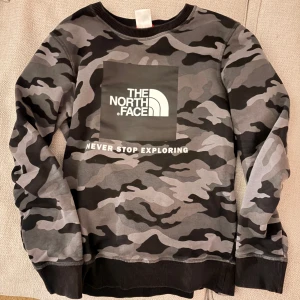 Camouflage tröja från The North Face - Säljer en cool camouflage tröja från The North Face med texten 'Never Stop Exploring' på framsidan. Tröjan har långa ärmar och en rund halsringning. Perfekt för den äventyrslystne!