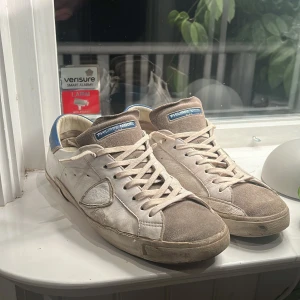 Vita sneakers från Philippe Model - Snygga vita sneakers från Philippe Model med blå detaljer på hälen och gråa partier på tån. Skorna har klassisk snörning och en stilren design. Sällsynt modell! Perfekta för en avslappnad och trendig look. Använt skick men inga defekter förutom att färgen lossnat lite på sidan och hälen. Hör av er vid frågor, priset är inte hugget i sten!