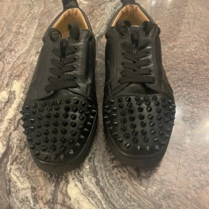 Svarta sneakers med nitar - Snygga svarta sneakers i skinn med nitar på tån och den ikoniska röda sulan. Skorna har snörning och en stilren design som sticker ut. Perfekta för dig som vill ha en edgy look.