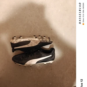 Svarta fotbollsskor från Puma - Säljer ett par svarta fotbollsskor från Puma med vita detaljer. Skorna har snörning och är perfekta för fotbollsträning. De är i storlek 40 och har en klassisk design med dobbar för bra grepp på planen.