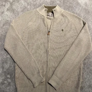 Beige stickad tröja från Morris - Säljer en stilren beige stickad kofta från Morris med dragkedja och ribbade detaljer. Perfekt för kyligare dagar. Den har en liten broderad logga på bröstet och långa ärmar.