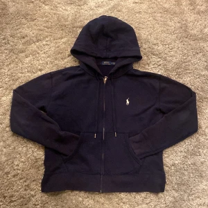 Ralph Lauren zip hoodie - Säljer en snygg marinblå zip hoodie från Polo Ralph Lauren. Den är i bra skick 8/10 kan vara någon enstaka fläck men inget man tänker på. Tröjan har slits där nere på sidorna som kanske inte syns på bilderna. Skriv gärna över frågor och funderingar, priset kan diskuteras.