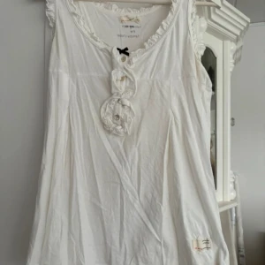 Vit ärmlös topp från Odd Molly - Säljer en söt vit ärmlös topp från Odd Molly med volangdetaljer runt halsen och en liten svart rosett framtill. Toppen har en knappdetalj och är perfekt för en avslappnad stil. Passar bra till både jeans och kjol. Storlek M - 3 