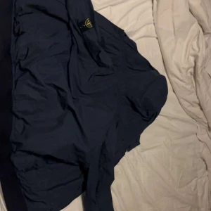 Mörkblå jacka från Stone Island Junior - Säljer en snygg mörkblå jacka från Stone Island Junior. Jackan har en klassisk design med huva och dragkedja. Perfekt för att hålla stilen i alla väder. Jackan är helt äkta. Kan sänka pris vid snabb affär.