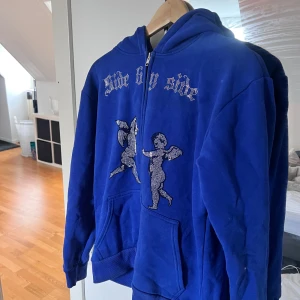 Side by Side Zip hoodie - Säljer en blå hoodie med dragkedja och ett unikt tryck av två änglar på framsidan. Texten 'Side by side' är tryckt ovanför i ett stilfullt typsnitt. Perfekt för en avslappnad stil. Det är rhinestone material och kommer från företaget side by side