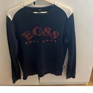 Svart tröja från Hugo Boss - Snygg svart tröja från Hugo Boss med vita axelpartier och röd text på framsidan. Perfekt för en stilren look. Tröjan har långa ärmar och rund halsringning.