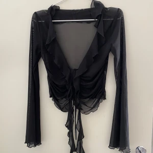 Svart transparent blus från Gina Tricot - Snygg svart transparent blus från Gina Tricot med långa ärmar och volangdetaljer. Blusen har en knytning framtill och en djup v-ringning.