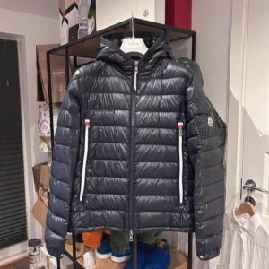 Svart pufferjacka från Moncler - Snygg svart pufferjacka från Moncler med huva och dragkedja framtill. Jackan har två framfickor med vita dragkedjor och en ärmficka med Moncler-logga. Perfekt för kyliga dagar.