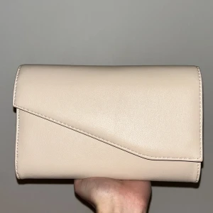 Beige kuvertväska med kedja - Elegant beige kuvertväska med en stilren design. Väskan har en avtagbar kedja som gör den lätt att bära både i handen och över axeln. Perfekt för att ge en sofistikerad touch till din outfit. 