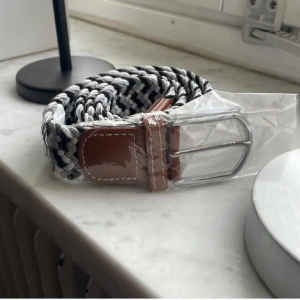 Flätat bälte - Snyggt flätat bälte i svart och vitt med bruna läderdetaljer och ett silverfärgat spänne. Perfekt för att ge en stilren touch till din outfit. 105 cm långt och passar alla då det är väldigt elastiskt.