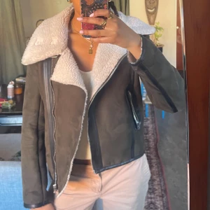 Brun Mockajacka med Vit Fuskpäls - Säljer en snygg brun/grön mockajacka med vit fuskpäls på kragen. Jackan har dragkedja och coola detaljer med knappar vid axlarna.