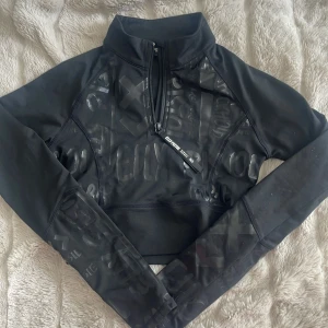 Svart croppad tröja från Elly Pistol - Snygg svart croppad tröja från Elly Pistol med glansigt mönster och halv dragkedja framtill. Perfekt för en sportig look med långa ärmar och en cool design. 