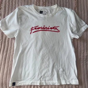 Printed tee - Säljer en vit t-shirt från Dedicated med röd text 'Feminista' på framsidan. Gjord av 100% ekologisk och Fairtrade-certifierad bomull. Perfekt för en avslappnad stil.